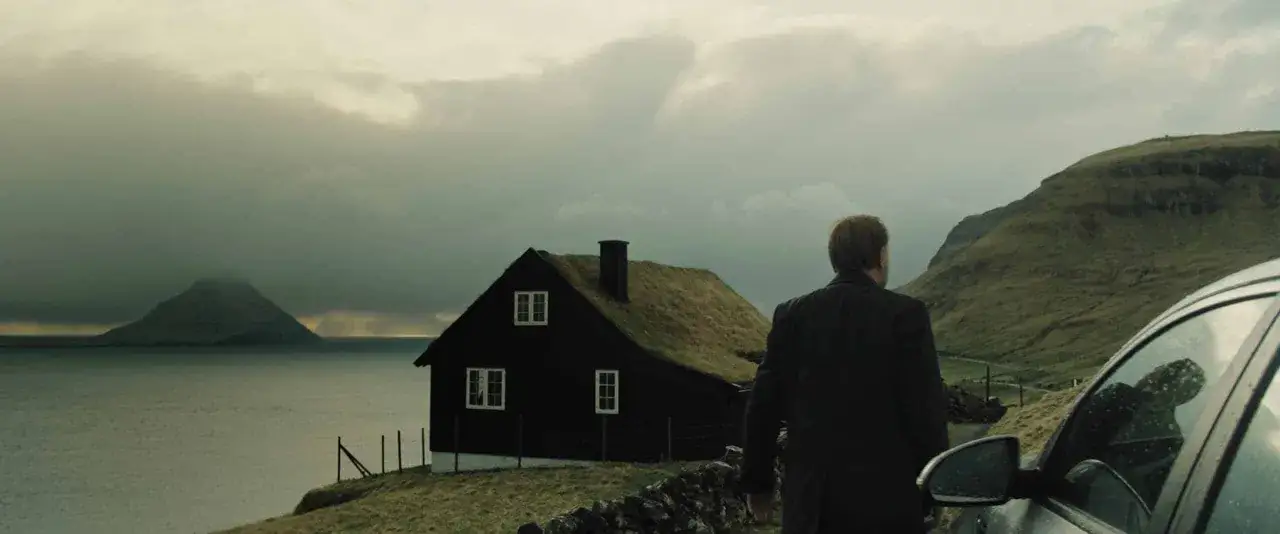 Man i kostym vid en bil, framför ett mörkt hus med grästak. I bakgrunden syns en bergig ö och en dramatisk himmel, en scen från en trom serie.