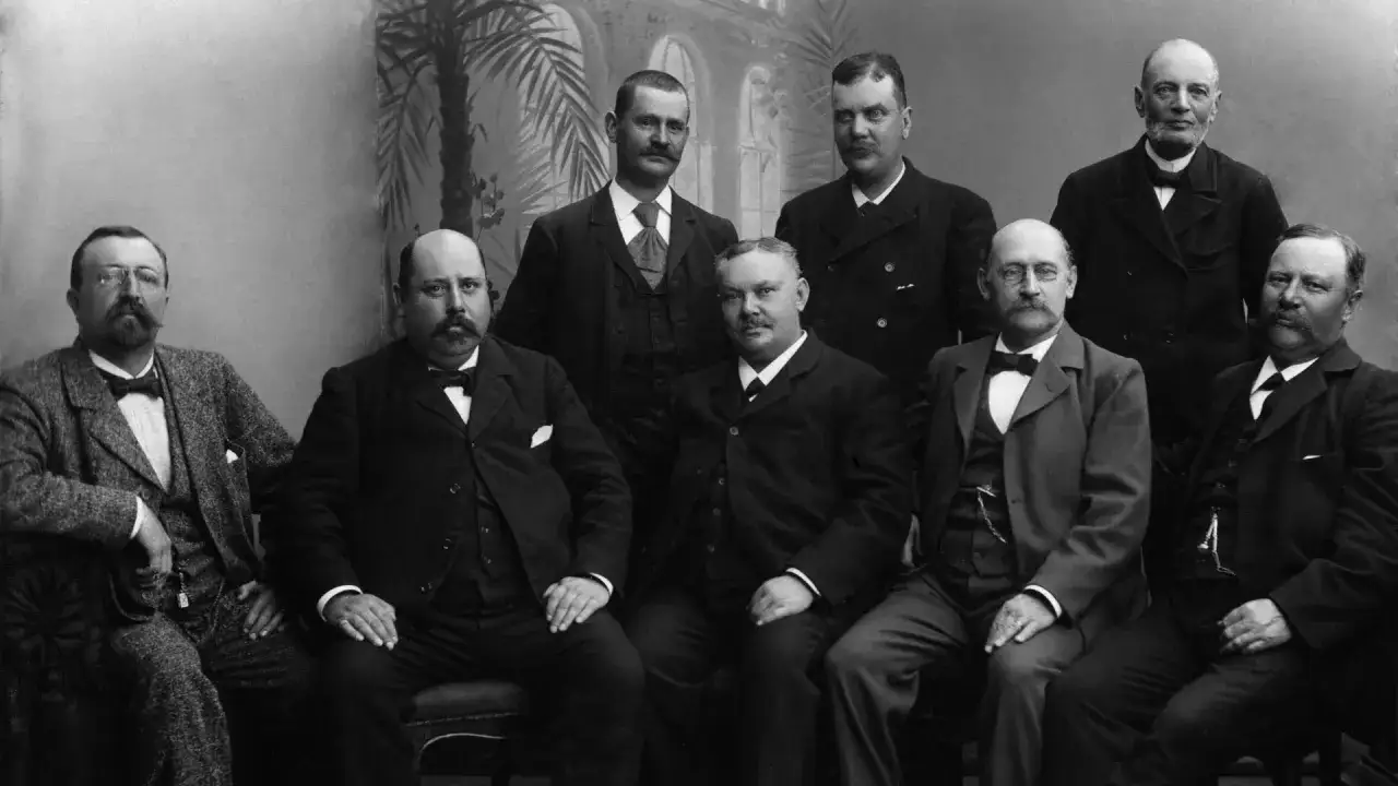 Gruppbild på män med mustasch och 1900-talets kläder, inklusive kostymer och flugor.