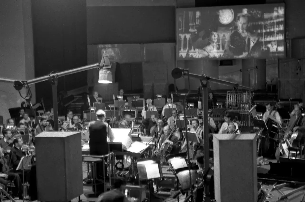 Stor orkester spelar film musik framför en bioduk med en filmscen. Dirigenten leder musikerna.