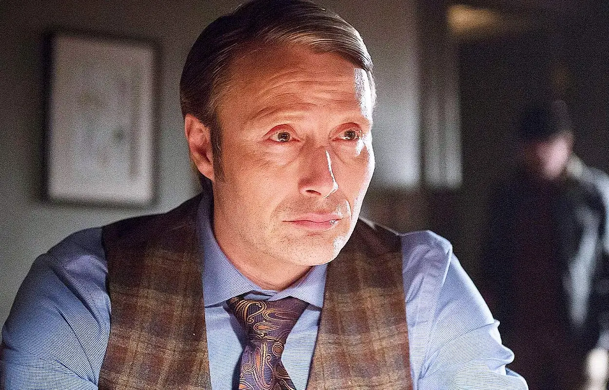 Mads Mikkelsen i en mörk scen från en serie. Han bär en rutig väst över en blå skjorta och en mönstrad slips.