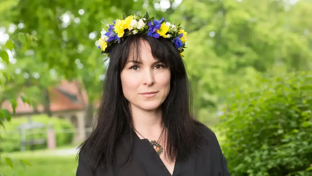 Nina Hemmingsson bär en blomsterkrans i gult och blått. Hon har mörkt hår och en svart klänning.