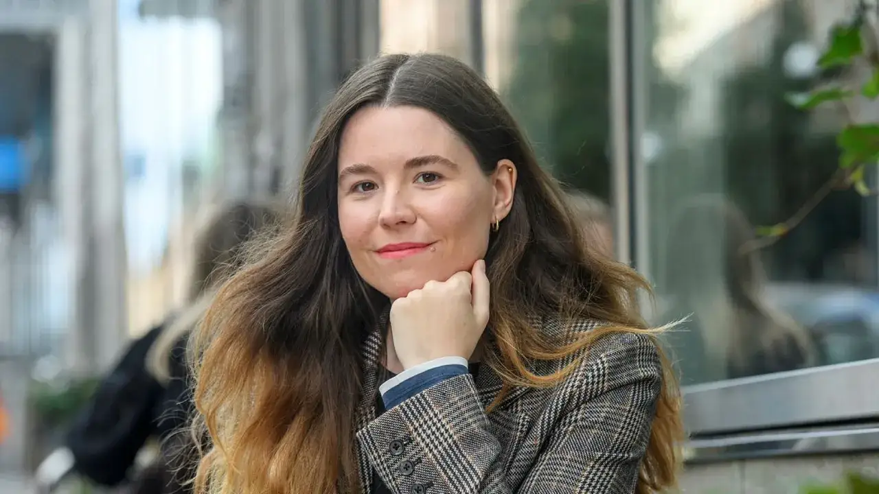 Johanna Hedman, med sitt långa, vågiga hår och rutiga kavaj, ler mot betraktaren.