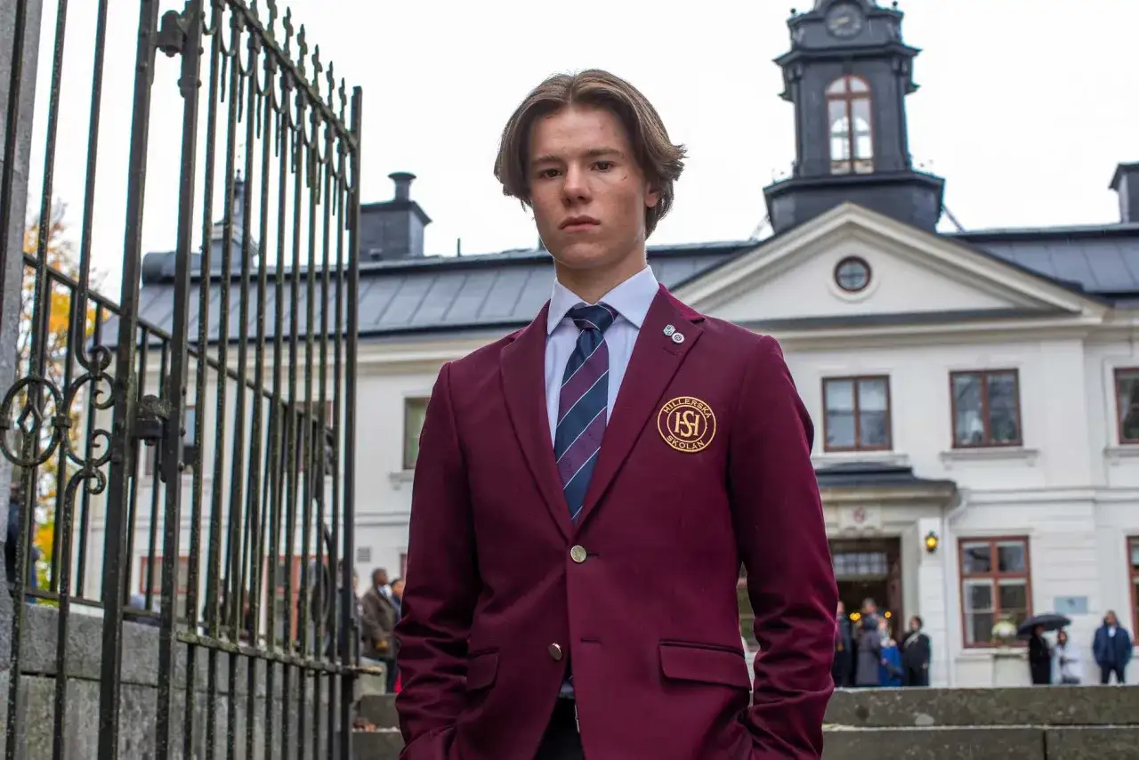 En ung man i skoluniform, som påminner om unga kungligheter, står framför en ståtlig byggnad.