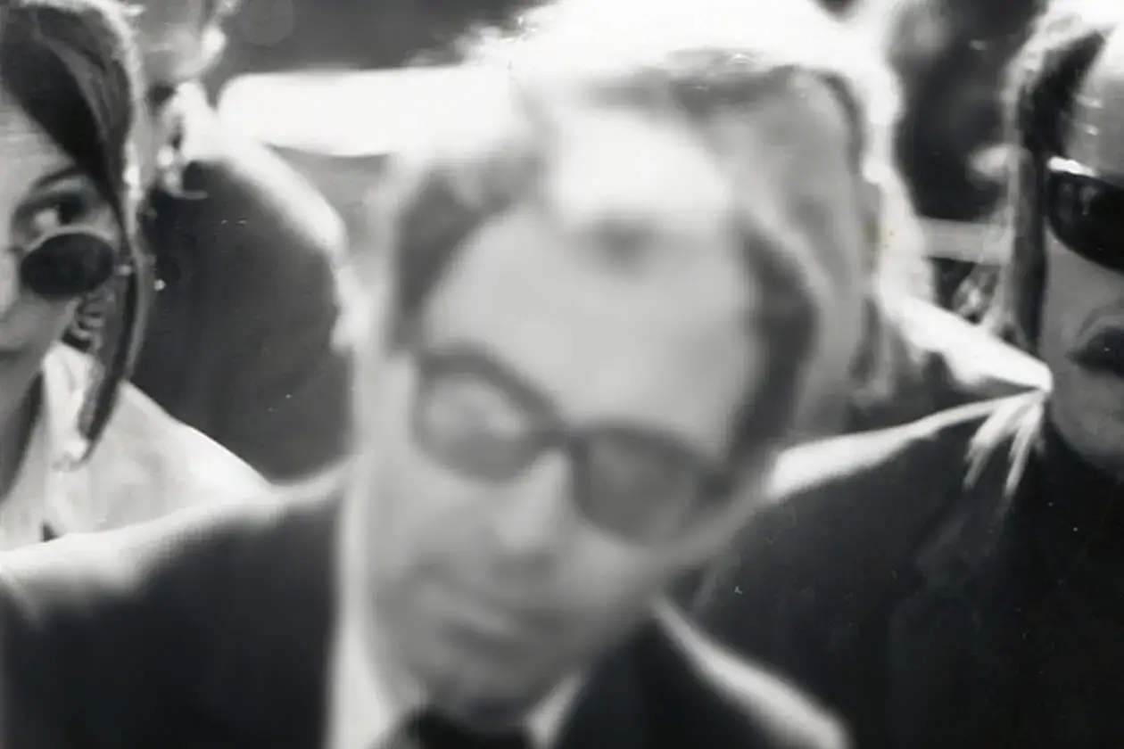 En suddig bild visar en grupp människor. I förgrunden syns en man med glasögon, möjligen Jean-Luc Godard, med en kvinna till vänster och en annan person till höger.