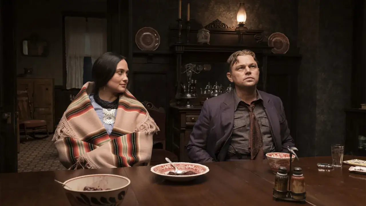 En kvinna i en traditionell filt och en man sitter vid ett bord. En scen fr&aring;n 