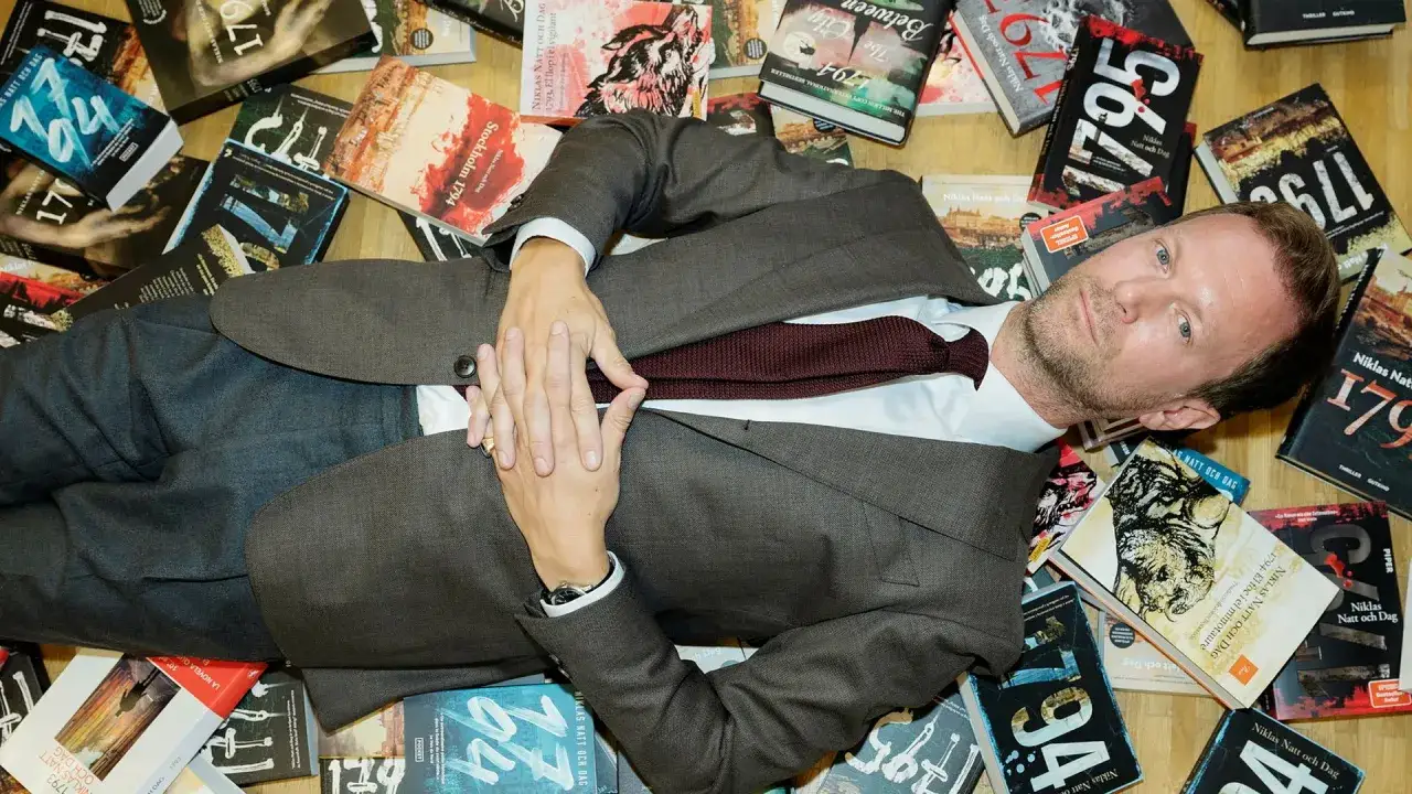 Man i kostym ligger omgiven av böcker av Niklas Natt och Dag.