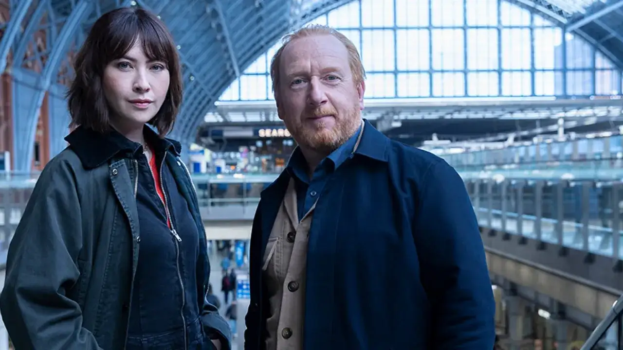 Två detektiver, en kvinna och en man, står i en stor, modern byggnad. De ser ut att vara på ett uppdrag som Chelsea detective.