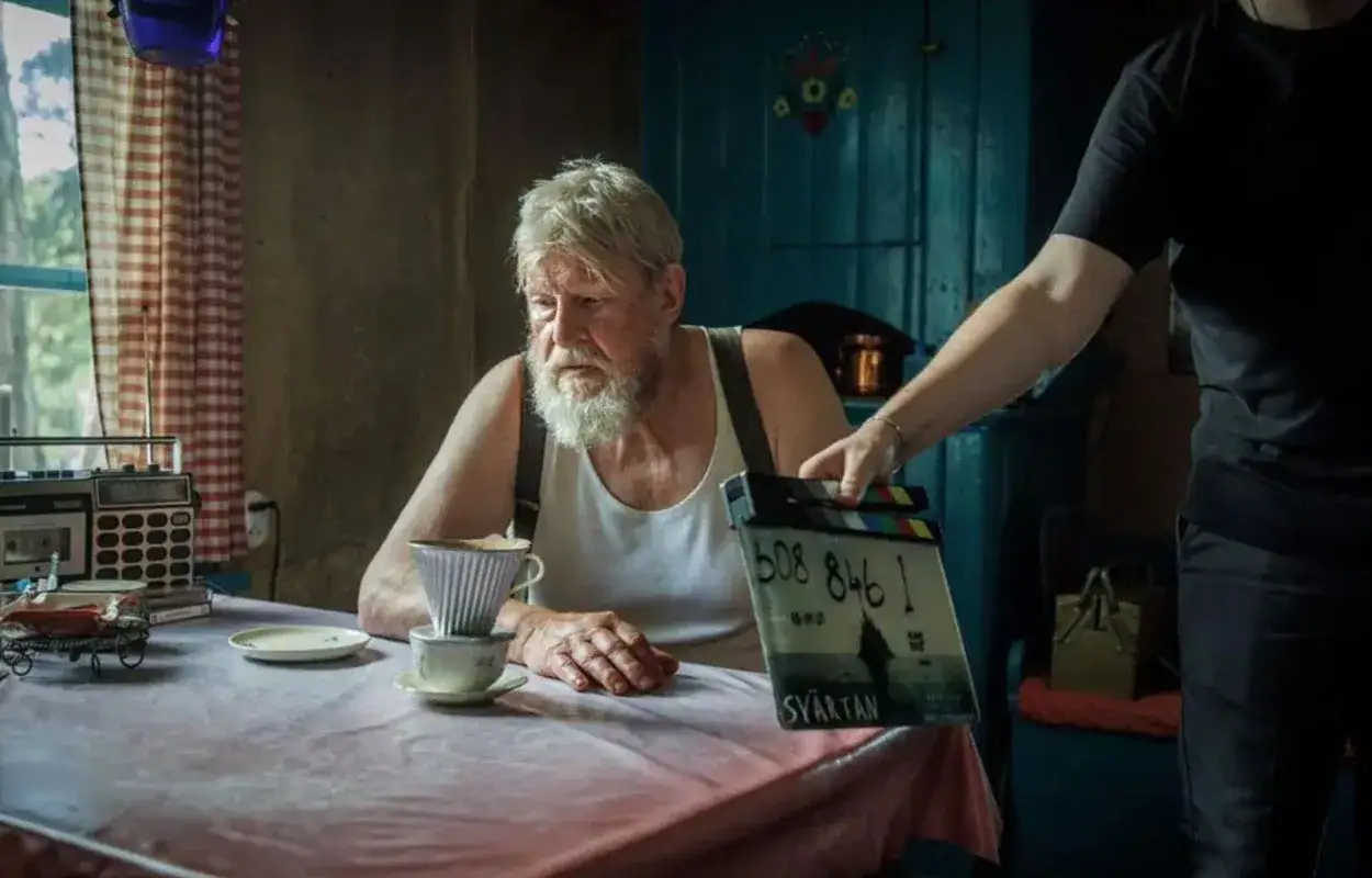 Rolf Lassgård sitter vid ett bord med en kaffebryggare. En person håller upp en filmklappa med texten
